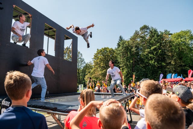 Acrobat flying over the trampoline beside the wall in a park with children watching in the foreground