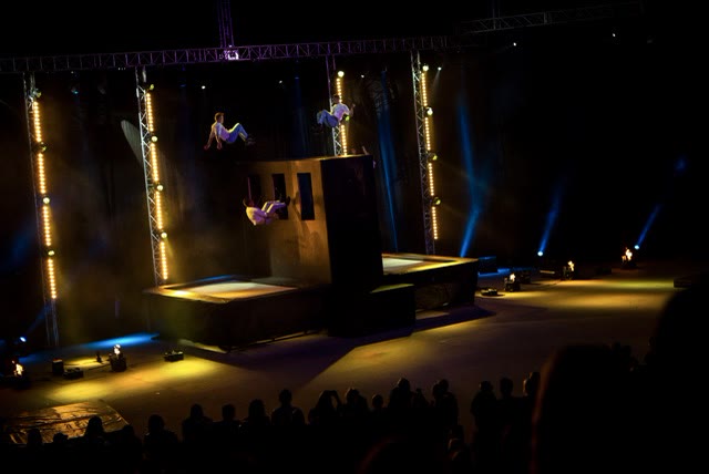 Wide view of acrobats on and around the trampoline wall on a dark stage with trusses and lights