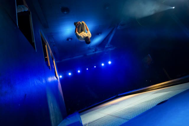 Acrobat upside down above the trampoline bed in blue stage light