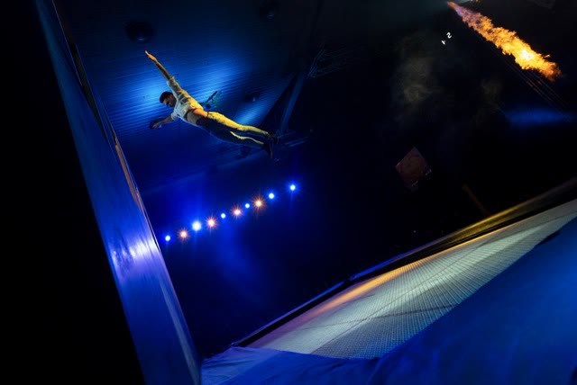 Acrobat flying horizontally beside the wall in blue light with a flame jet in the background