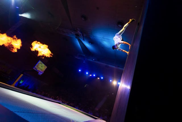 Acrobat in a handstand on the trampoline wall with two flame effects and an audience below