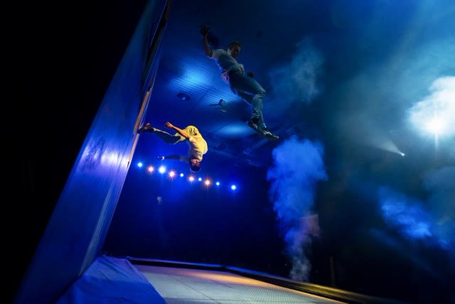 Two acrobats flipping beside the blue wall amid stage smoke