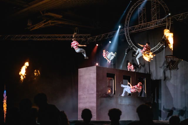 Acrobats in red patterned shirts leaping off the trampoline wall with a metal ring and flames on a large stage