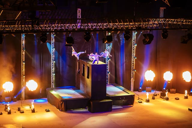 Top view of acrobats flying off the trampoline wall with several flame jets on the stage