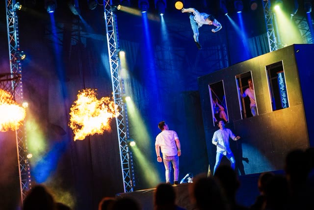 Acrobat flying from the trampoline wall while two performers watch from below, with a flame on the left