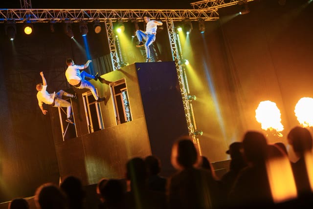 Acrobats jumping off the trampoline wall in warm stage light with flames and audience silhouettes