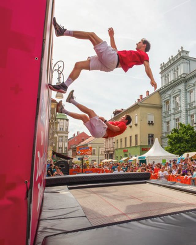 Two acrobats flipping beside the pink trampoline wall in front of a large crowd on a town square