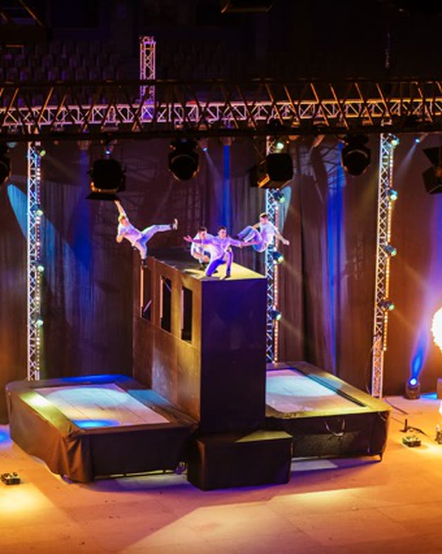 Acrobats on and around a two-sided trampoline wall on a stage with flame effects, seen from above